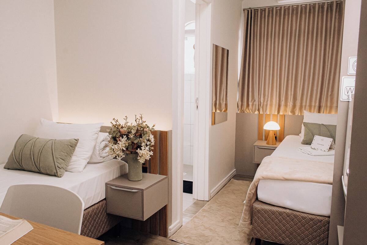 Una habitación de hotel con dos camas, una mesita de noche con lámpara, un florero con flores y una pequeña mesa con una silla.