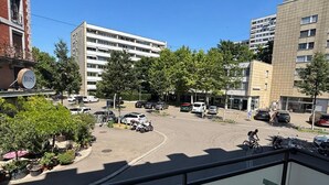 Vista desde un balcón sobre un estacionamiento con varios coches, un edificio con un letrero que dice 'ROS' y un cielo azul despejado.