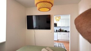 Un dormitorio con cama, televisor montado en la pared y cocina visible a través de un vano abierto.