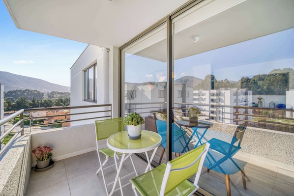 A balcony with a table and chairs, a potted plant, and a view of buildings and mountains.