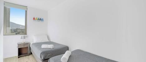 A bedroom with two beds, a nightstand, a lamp, and a window with a view.