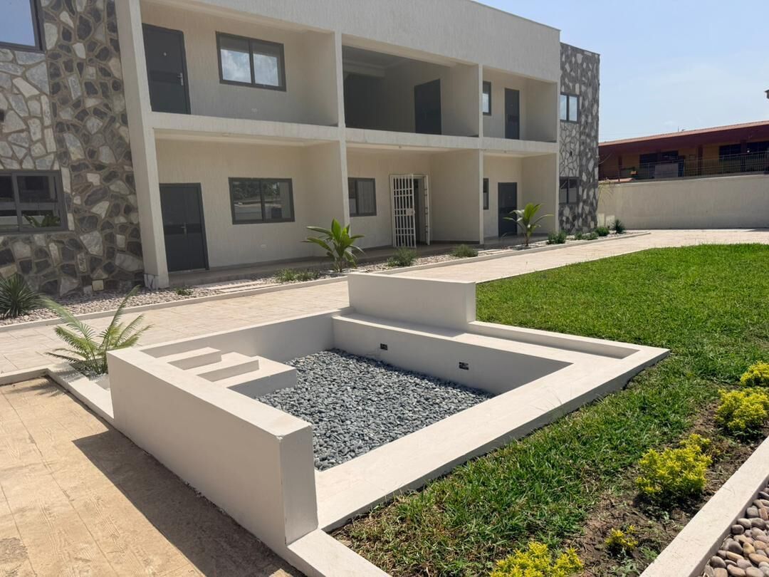 Un bâtiment moderne à plusieurs étages, doté d’une façade en pierre, d’un jardin aménagé avec un bassin rectangulaire et surplombé d’un ciel dégagé.