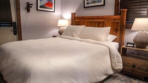 A neatly made bed with a wooden headboard, two bedside lamps, and a small table with a lamp. - Deluxe King One-Bedroom Cottage (Mobility Accessible) (Bronson)