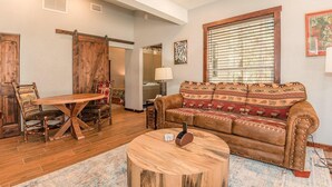 A cozy living room with a wooden dining table, a sofa with patterned cushions, and a large window with blinds. - Deluxe King One-Bedroom Cottage (Mobility Accessible) (Bronson)