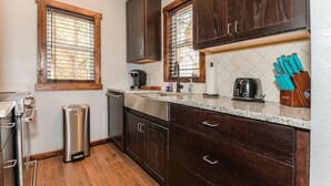 A kitchen with dark wooden cabinets, a stainless steel range hood, a coffee maker, a toaster, and a trash can. - Deluxe King One-Bedroom Cottage (Mobility Accessible) (Bronson)