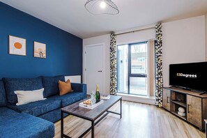 Ein modernes Wohnzimmer mit einem blauen Sofa, einem Couchtisch mit Flasche und Gläsern, einem Flachbildfernseher und Blick durch eine Glastür.