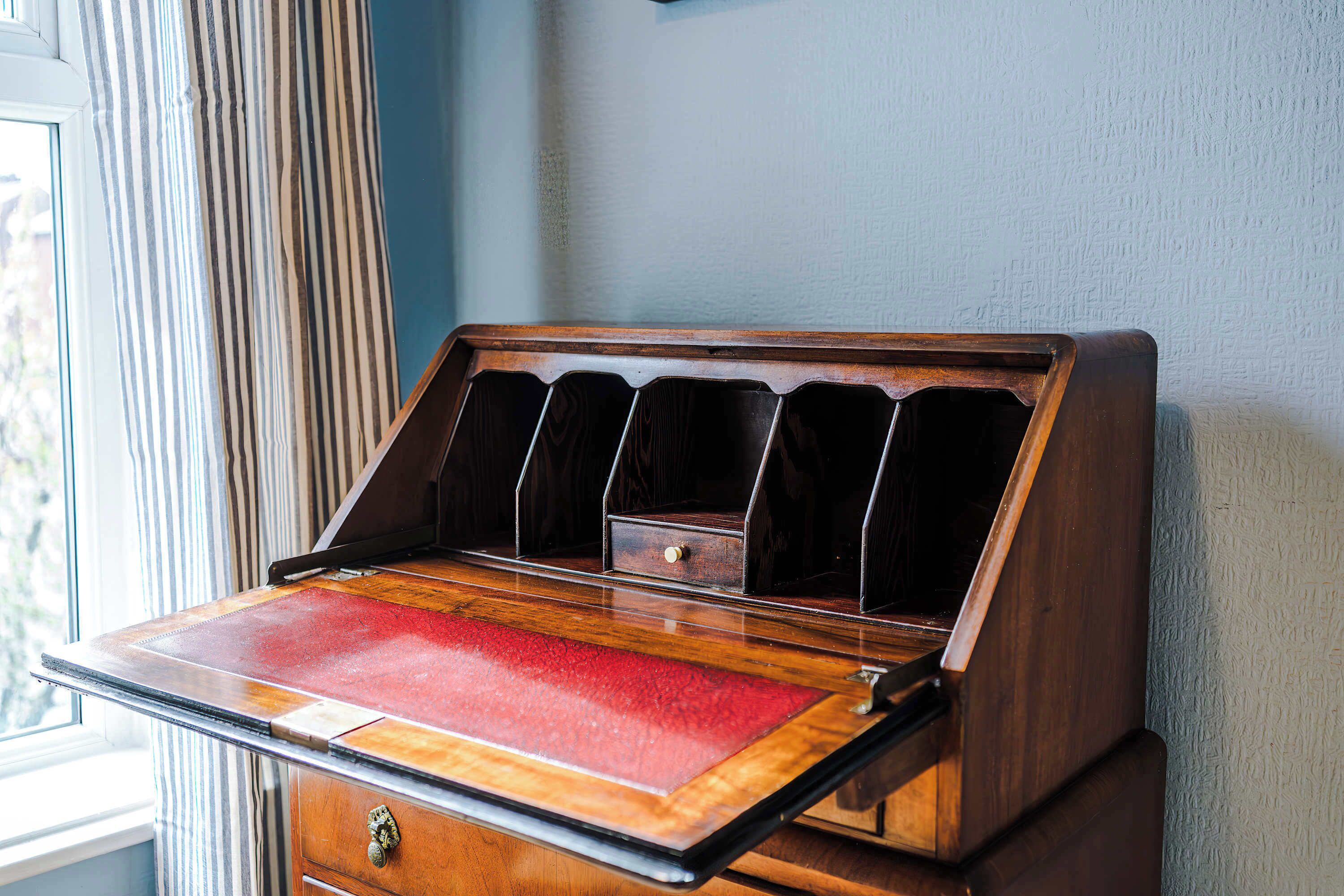 Una scrivania in legno con piano di lavoro rosso, un cassetto con pomello e una finestra con tende a righe.