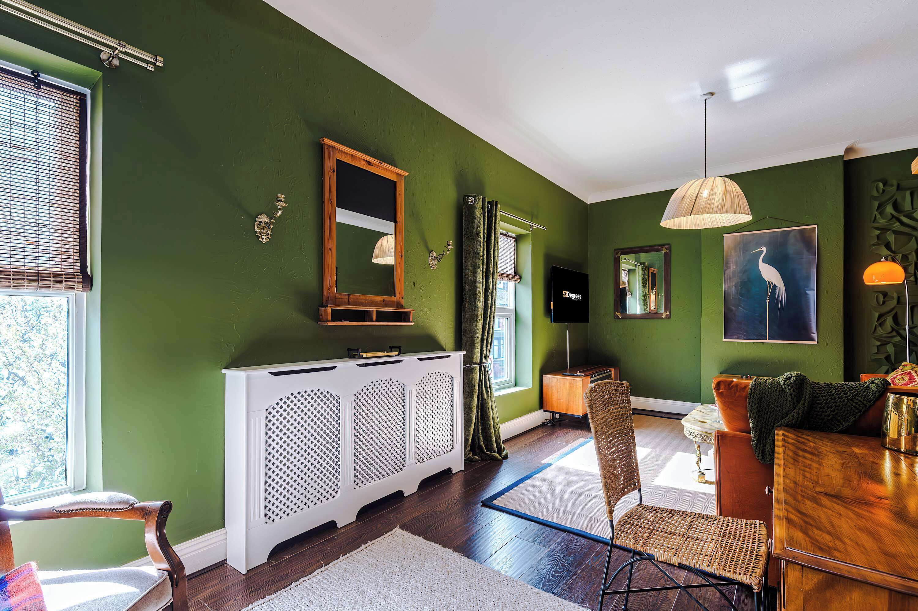 A room with a green wall, a white radiator, a wooden cabinet, a dining table with chairs, and a television.