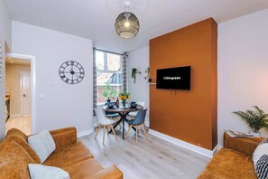 A modern living room with a dining area, a large clock, and a flat-screen TV mounted on the wall. - Perfect Modern 4 Bed in Crewe, Great for Business (Cheshire East)