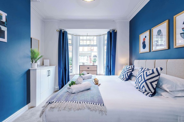 Contemporary comfort: a large bed with white duvet, blue-patterned pillows, folded towels, and warm lighting. Framed abstract art on a deep blue wall enhances the stylish, serene vibe.
