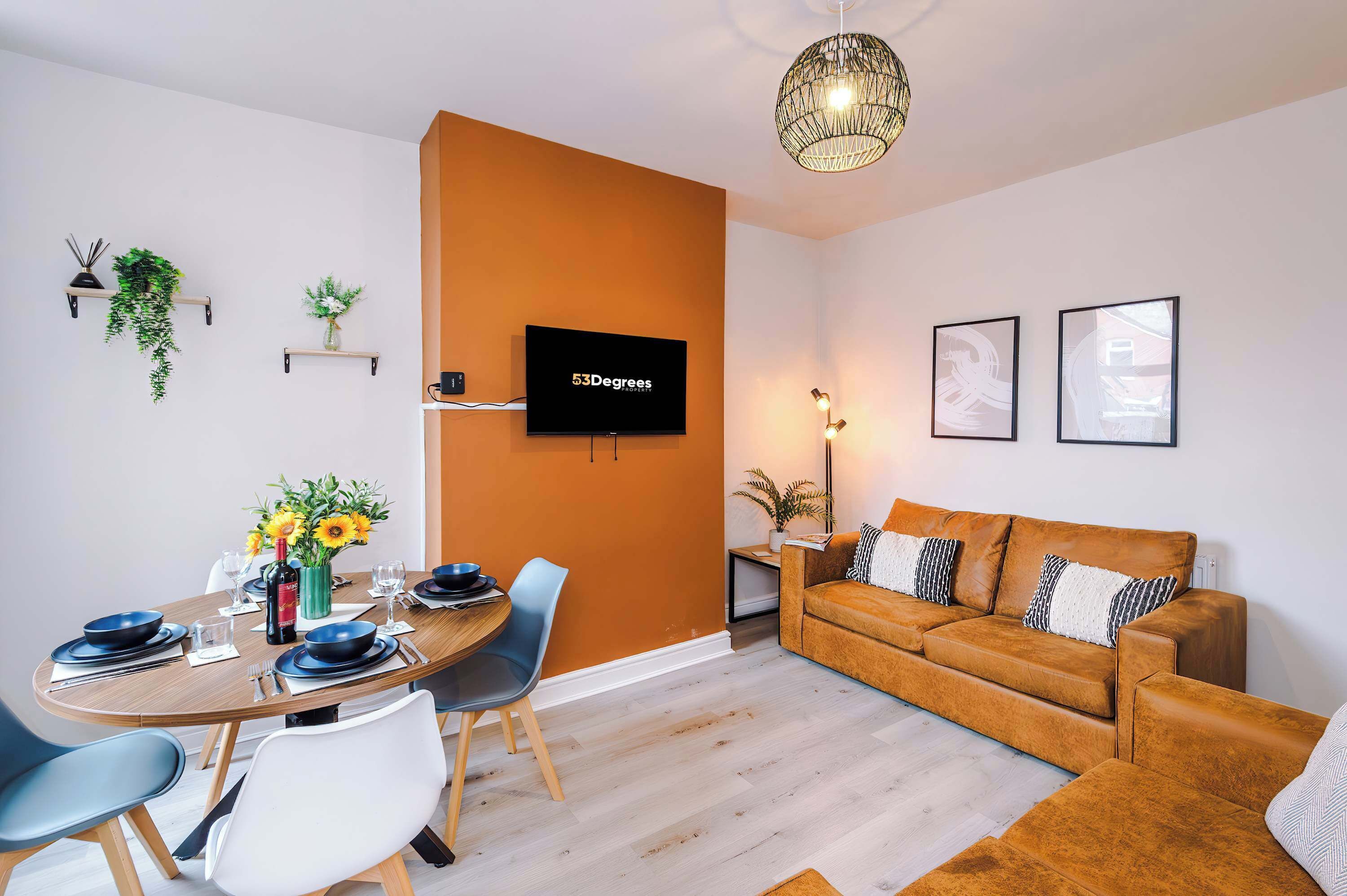 A chic open-plan space featuring a burnt orange accent wall, mounted TV, tan leather sofas, patterned cushions, framed art, and a round dining table set with wine and plates, enhanced by greenery and stylish lighting.