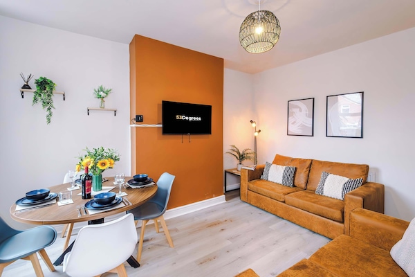 A chic open-plan space featuring a burnt orange accent wall, mounted TV, tan leather sofas, patterned cushions, framed art, and a round dining table set with wine and plates, enhanced by greenery and stylish lighting.