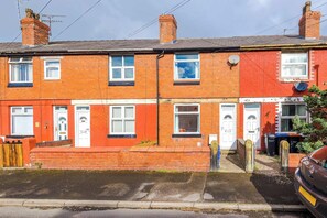 A red brick terraced house with two visible front doors, a small front garden, and a parked car. - Great 3-Bed Home, Perfect for Work & Contractors! (Cheshire West and Chester)
