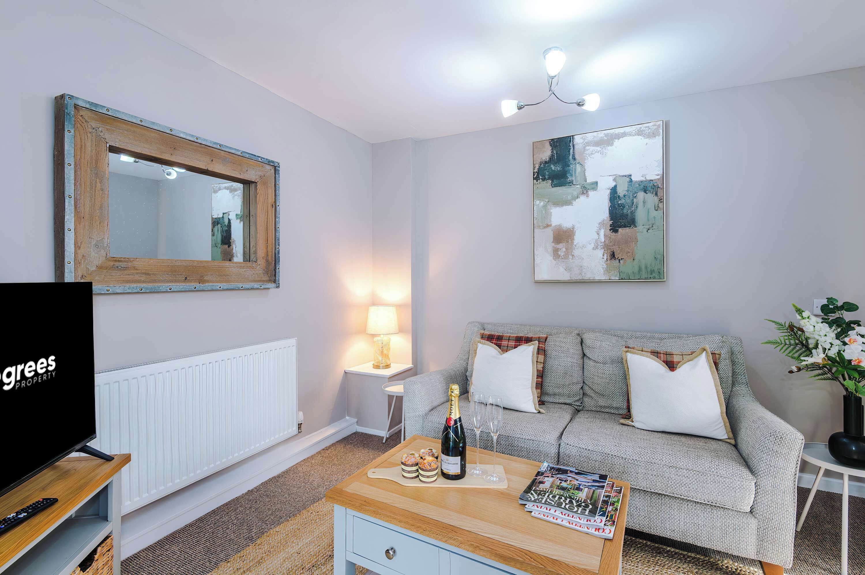 A cozy modern living room with a gray sofa, patterned throw, and champagne on the coffee table. Abstract art and a wooden-framed mirror add style, while soft lighting creates a warm, inviting atmosphere.