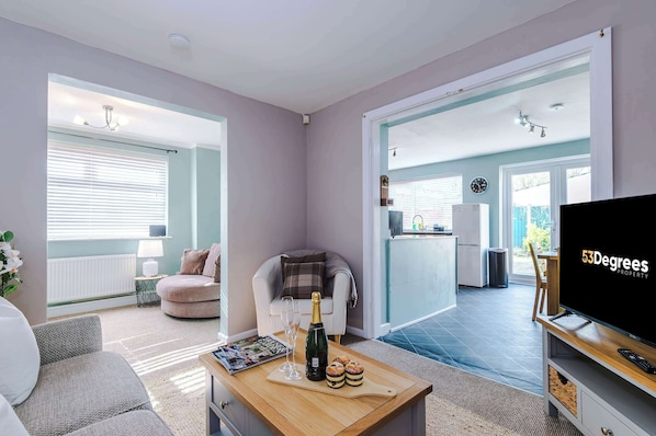 A living room with a sofa, a coffee table with a bottle and pastries, a TV, and a view into a kitchen and dining area. - Comfortable 4 Bed Home, Perfect for Business Stays (Elton)