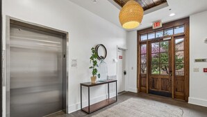 Un hall d’entrée moderne avec une porte en bois, une porte en verre, un ascenseur en métal, une plante en pot et un tapis à motifs.