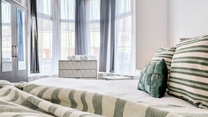 Ein Hotelzimmer mit Bett, Nachttisch, einem Fenster mit Vorhängen und Blick auf Gebäude.