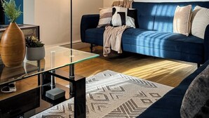 Ein Wohnzimmer mit blauem Sofa, Glastisch, Stehlampe und einem gemusterten Teppich.