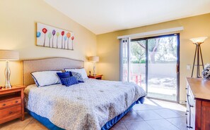 A bedroom with a bed, nightstands, a lamp, a painting, and a view through a sliding door. - Palm Desert Condo | Pool + Golf Nearby (Palm Desert)