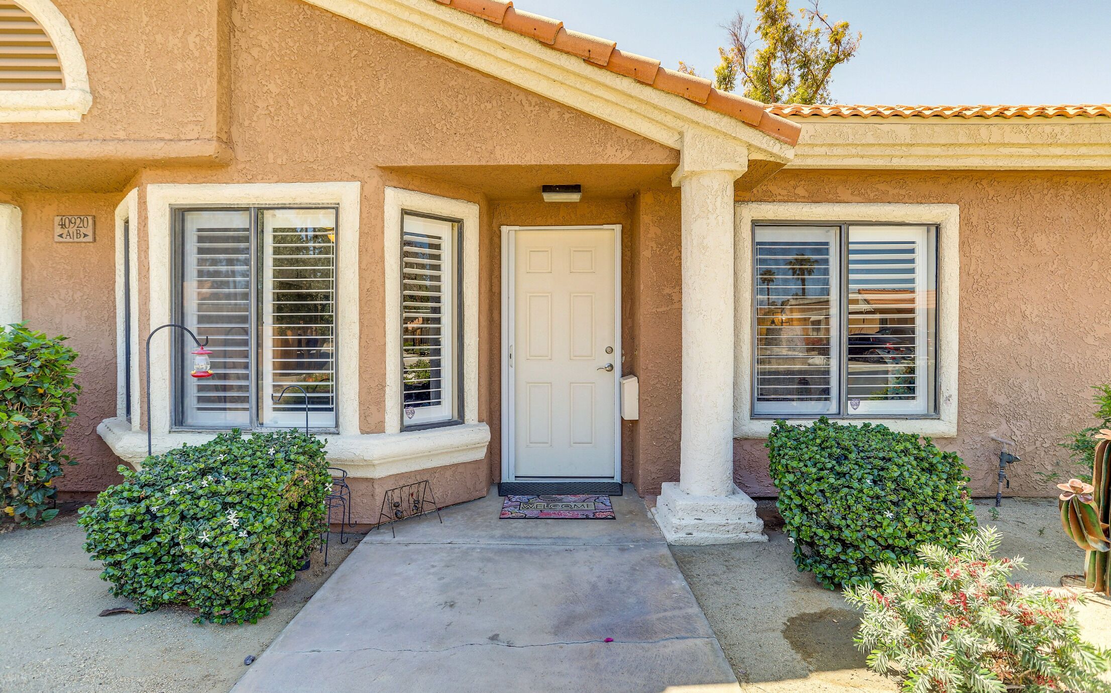 A house with a white door, two windows with blinds, a welcome mat, and a small garden with bushes.