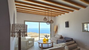 A modern living room with a high ceiling, wooden beams, a large window with a view, and a seating area with a sofa and a coffee table. - Atlas in Mykonos With 8 Bedrooms and 8 Bathrooms (Mykonos)