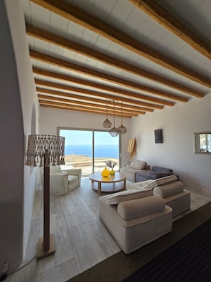 Ein modernes Wohnzimmer mit hoher Decke, Holzbalken, einem großen Fenster mit Aussicht und einer Sitzecke mit Sofa und Couchtisch.