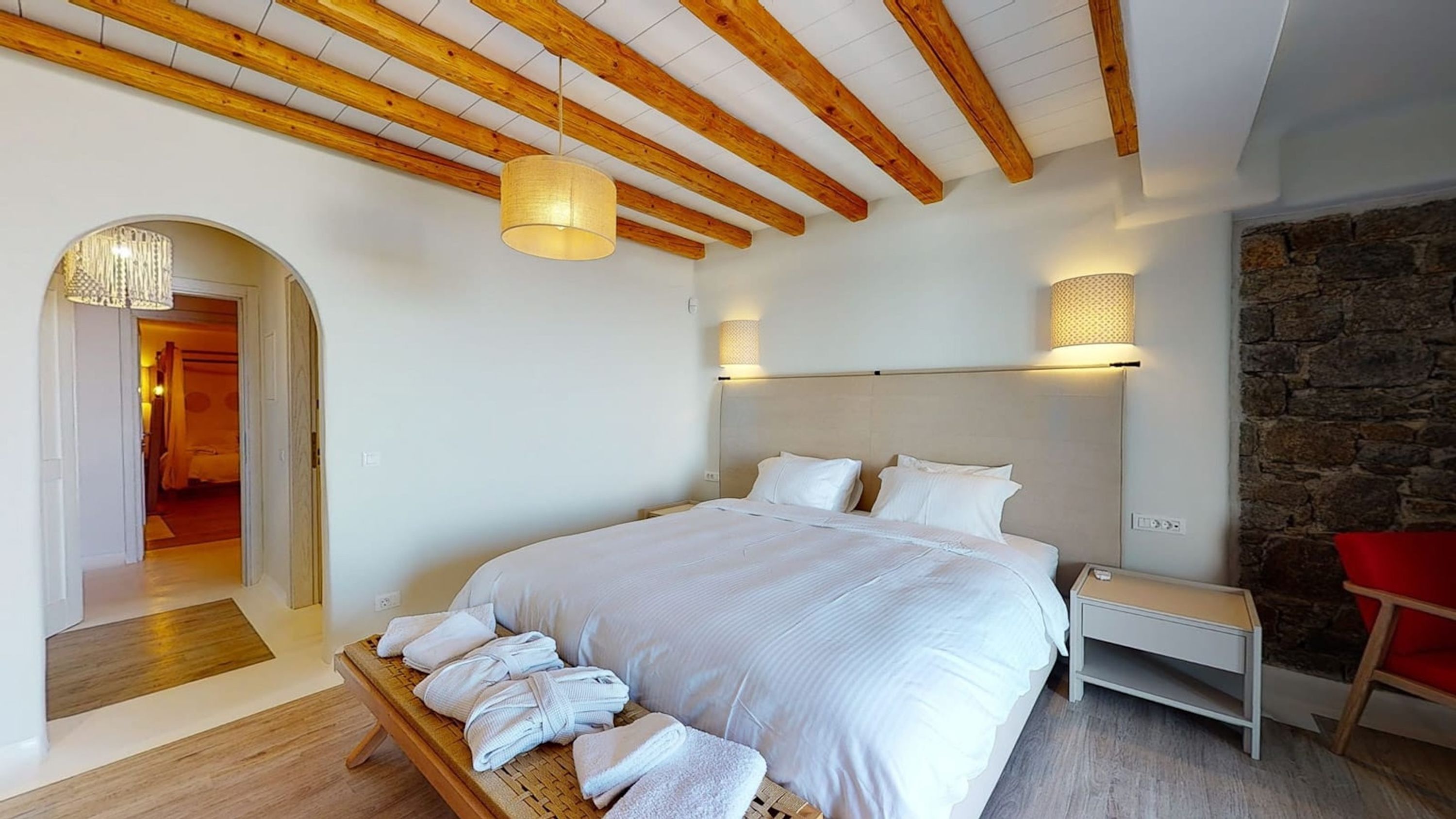 A bedroom with a large bed, wooden beams, a stone wall, and a red chair.
