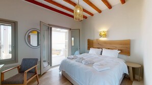 A bedroom with a large bed, a chair, a bedside table, a wall-mounted lamp, and a view through an open door. - Atlas in Mykonos With 8 Bedrooms and 8 Bathrooms (Mykonos)