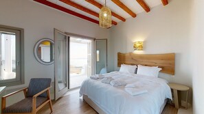 Ein Schlafzimmer mit einem großen Bett, einem Sessel, einem Nachttisch, einer Wandlampe und Blick durch eine offene Tür.
