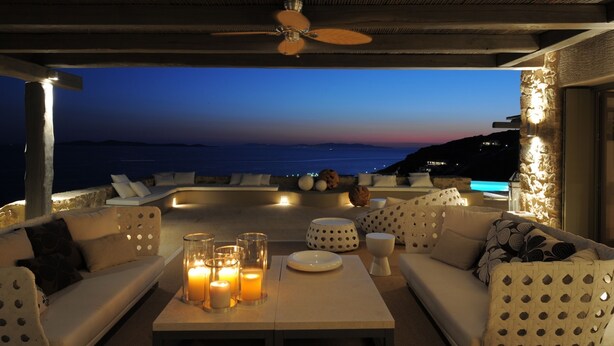Zona de estar al aire libre con vista al mar al anochecer, un ventilador de techo y una mesa con velas.