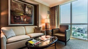 Una camera d'hotel con divano, poltrona, tavolino in vetro, un quadro appeso al muro, una lampada e un'ampia finestra con vista sulla città.