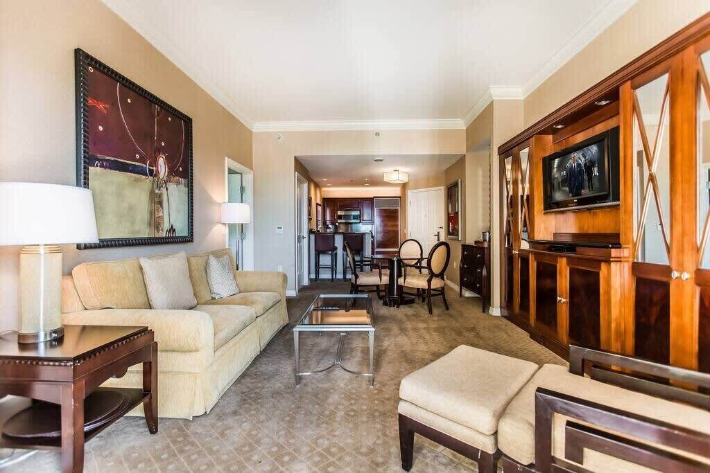 A hotel room with a sofa, a coffee table, a TV mounted on a wooden cabinet, and a dining area in the background.