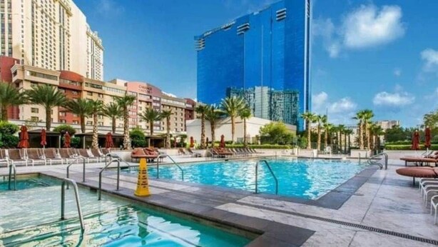 Zona de piscina de hotel con varias piscinas, palmeras y un edificio grande al fondo.