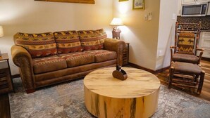 Una sala de estar con un sofá estampado, una mesa de centro redonda de madera, una lámpara y un sillón rústico.