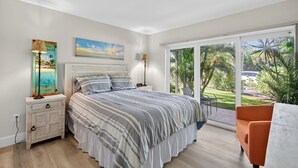 A bedroom with a bed, nightstands, a lamp, a painting, and a view of a garden through large windows. - Enjoyable 1-bedroom 1-bath condo in charming Marco Island. (Marco Island)