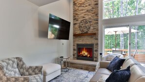 Un salon doté d’une cheminée en pierre, d’un téléviseur fixé au mur et d’une grande fenêtre donnant sur la forêt.