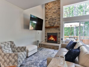 A living room with a stone fireplace, a mounted TV, and a large window with a view of trees. - Spacious 2 bdrm, 2 Bath with Loft, 1380 Sq Ft.  Many onsite amenities with Golf/Skiing near by! (Harbor Springs)