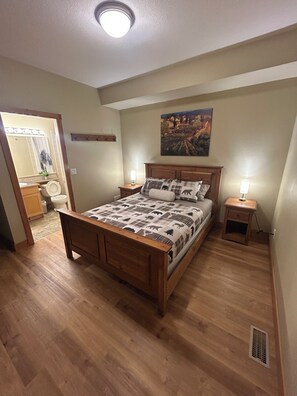 Une chambre à coucher avec un lit en bois, deux tables de chevet avec des lampes, un tableau au mur et une salle de bain visible à travers une porte ouverte.
