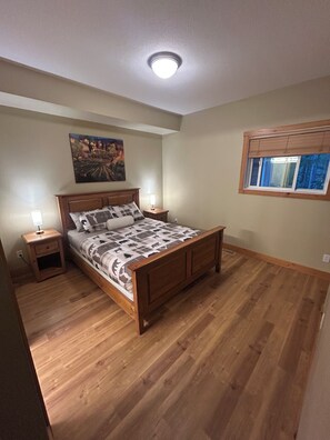 A bedroom with a wooden bed, a nightstand, a wall-mounted lamp, a window with blinds, and a ceiling light.