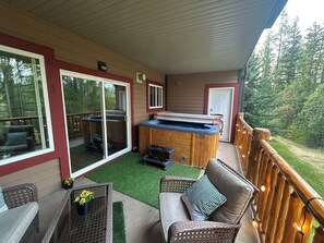 A covered deck with outdoor furniture, a hot tub, and a view of the surrounding forest.