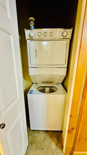 A compact laundry room with a stacked washer and dryer, a door, and a tiled floor. - Ski & Golf Getaway Suite - private hot tub - 2 bed (Kimberley)