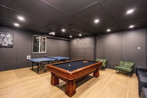 A pool table with a ball, a ping pong table, and a green cushioned chair in a room with wooden flooring and grey walls.