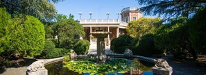 Ein Garten mit einem Brunnen, ein Gebäude mit Säulen und ein klarer Himmel.