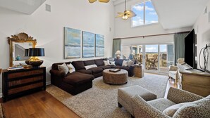 A spacious living room with a large sectional sofa, a round ottoman, and a coffee table. There are two pendant lights, a large mirror, and artwork on the walls. The room has a wooden floor and a large window with a view of the outdoors.