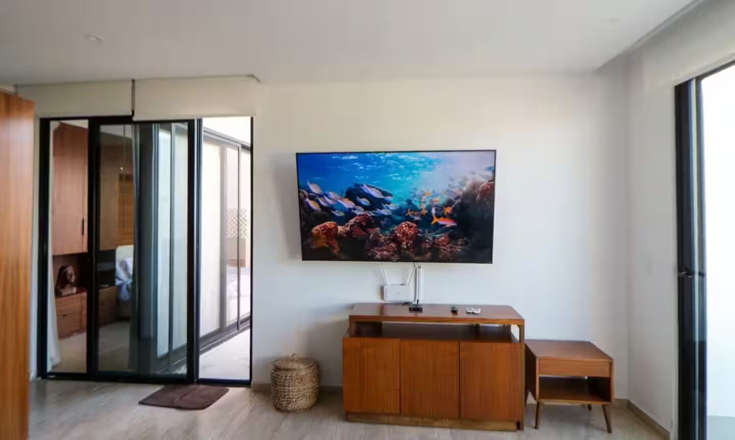 Un salon moderne doté d’un grand téléviseur à écran plat fixé au mur, d’un meuble de rangement en bois et d’une porte-fenêtre coulissante donnant sur une autre pièce.