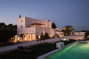 Una villa moderna con piscina, zona de estar exterior y un jardín bien iluminado al anochecer.