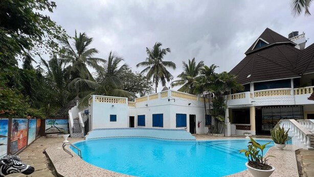 A swimming pool surrounded by a white building with blue windows, palm trees, and a stone pathway. - Luxurious 4-bedroom Holiday House With Pool Access in Diani Beach (Diani Beach)