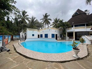 Una piscina rodeada por un edificio blanco con ventanas azules, palmeras y un sendero de piedra.