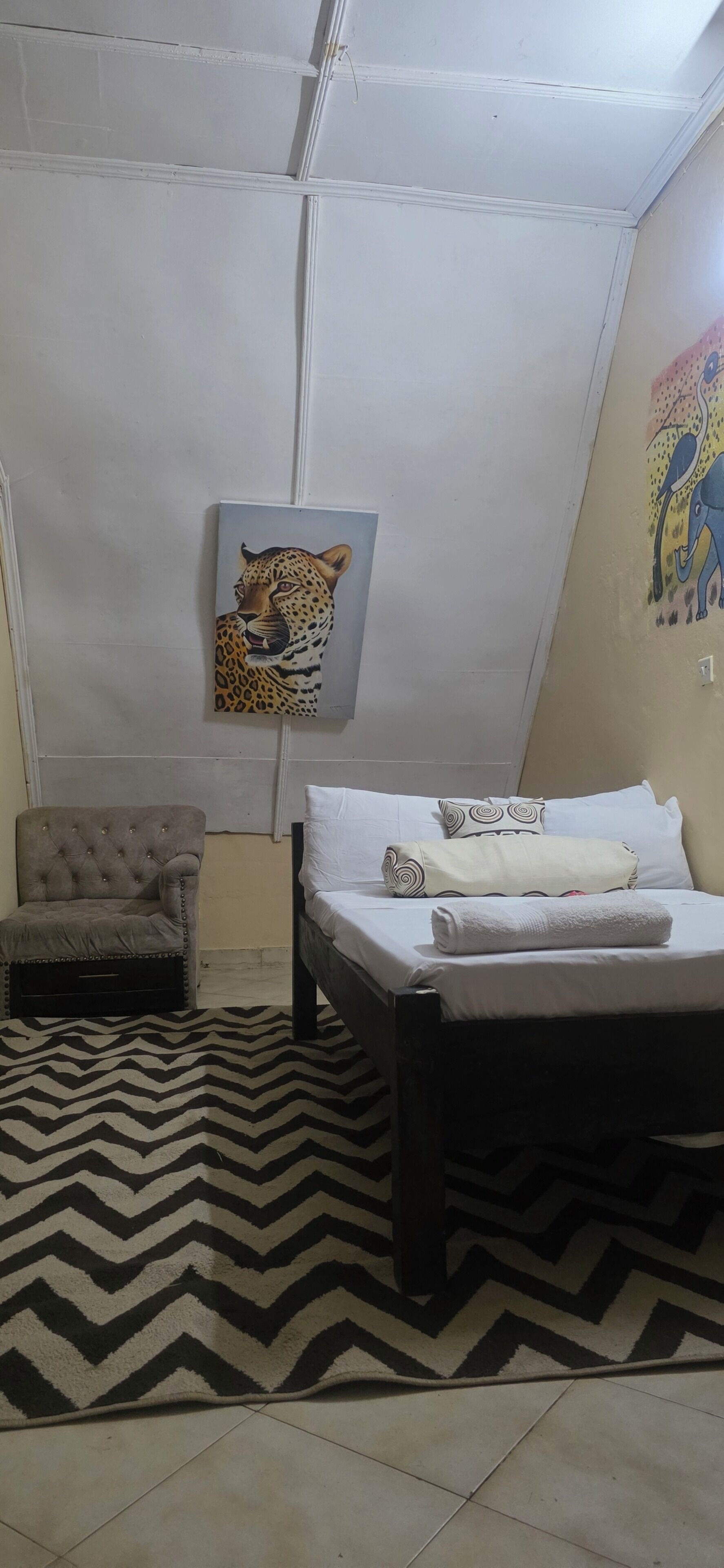 A room with a bed, a chair, a chevron-patterned rug, a leopard painting, and an elephant painting on the wall.