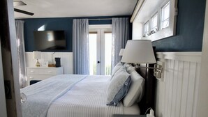 Ein Hotelzimmer mit einem Bett, einem an der Wand montierten Fernseher, einem Nachttisch mit Lampe, einem Telefon und einem Fenster mit Vorhängen.